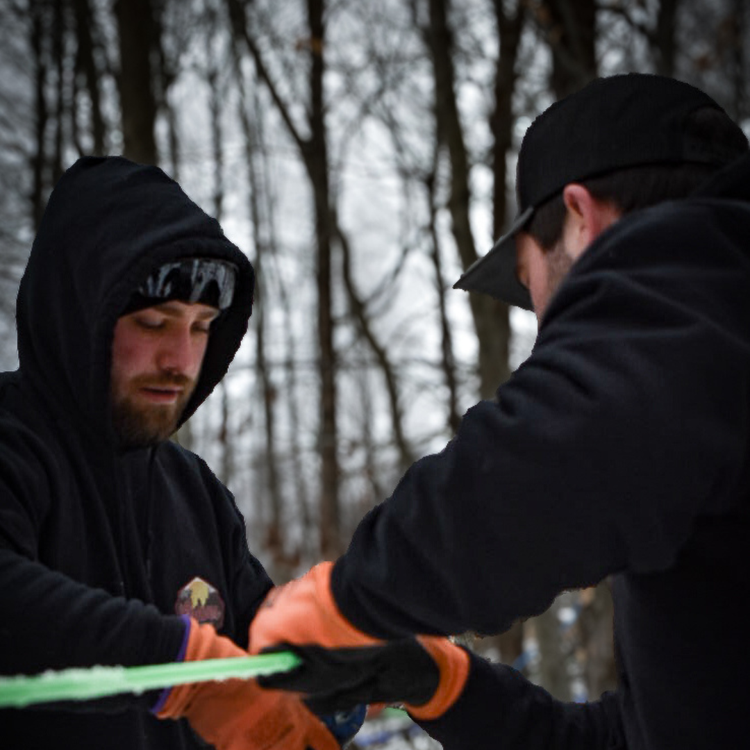 Two Graves Maple Products employees inspecting green sap lines in snowy Pennsylvania sugarbush during maple syrup season.