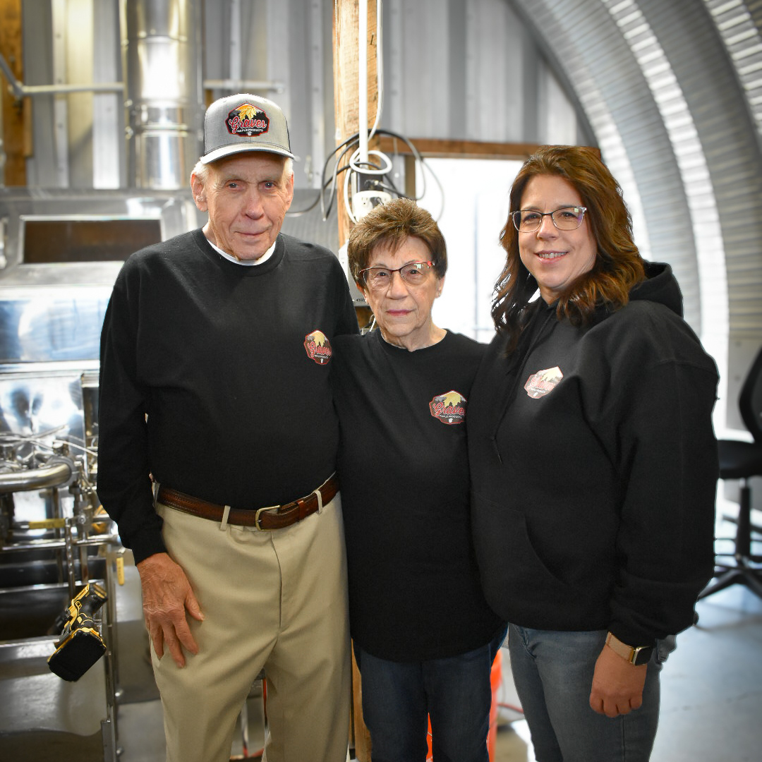 Graves Maple Products team members standing in front of stainless steel maple syrup processing equipment inside the sugarhouse, showcasing the production setup during maple season.