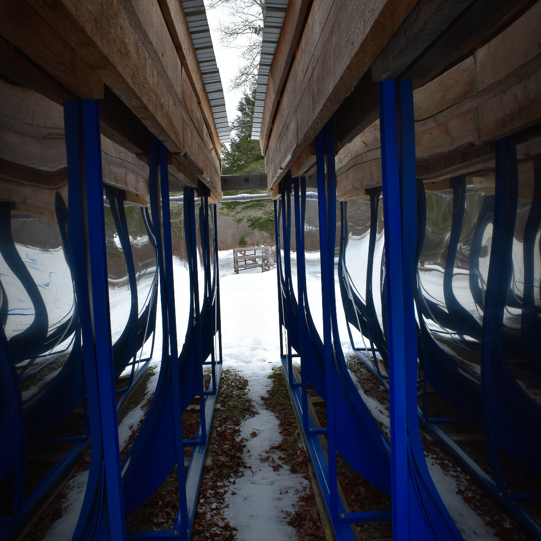 Stainless Steel Sap Holding Tanks in Snowy Sugarbush – Graves Maple Products
View between two rows of shiny stainless steel sap holding tanks mounted under wooden sheds at Graves Maple Products, with snow on the ground and a scenic backdrop of trees and a wooden gate.