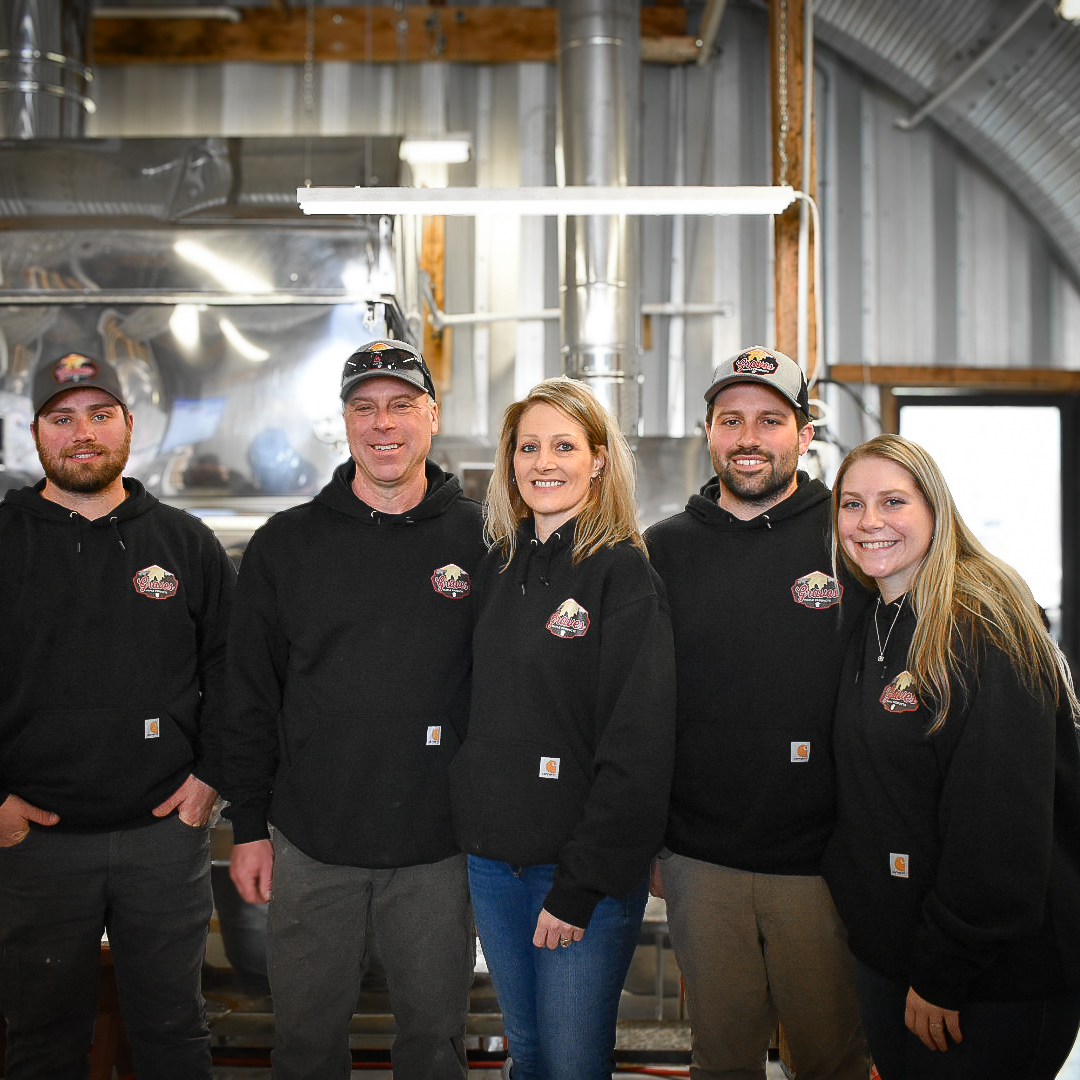 Graves Maple Products team members standing in front of stainless steel maple syrup processing equipment inside the sugarhouse, showcasing the production setup during maple season.