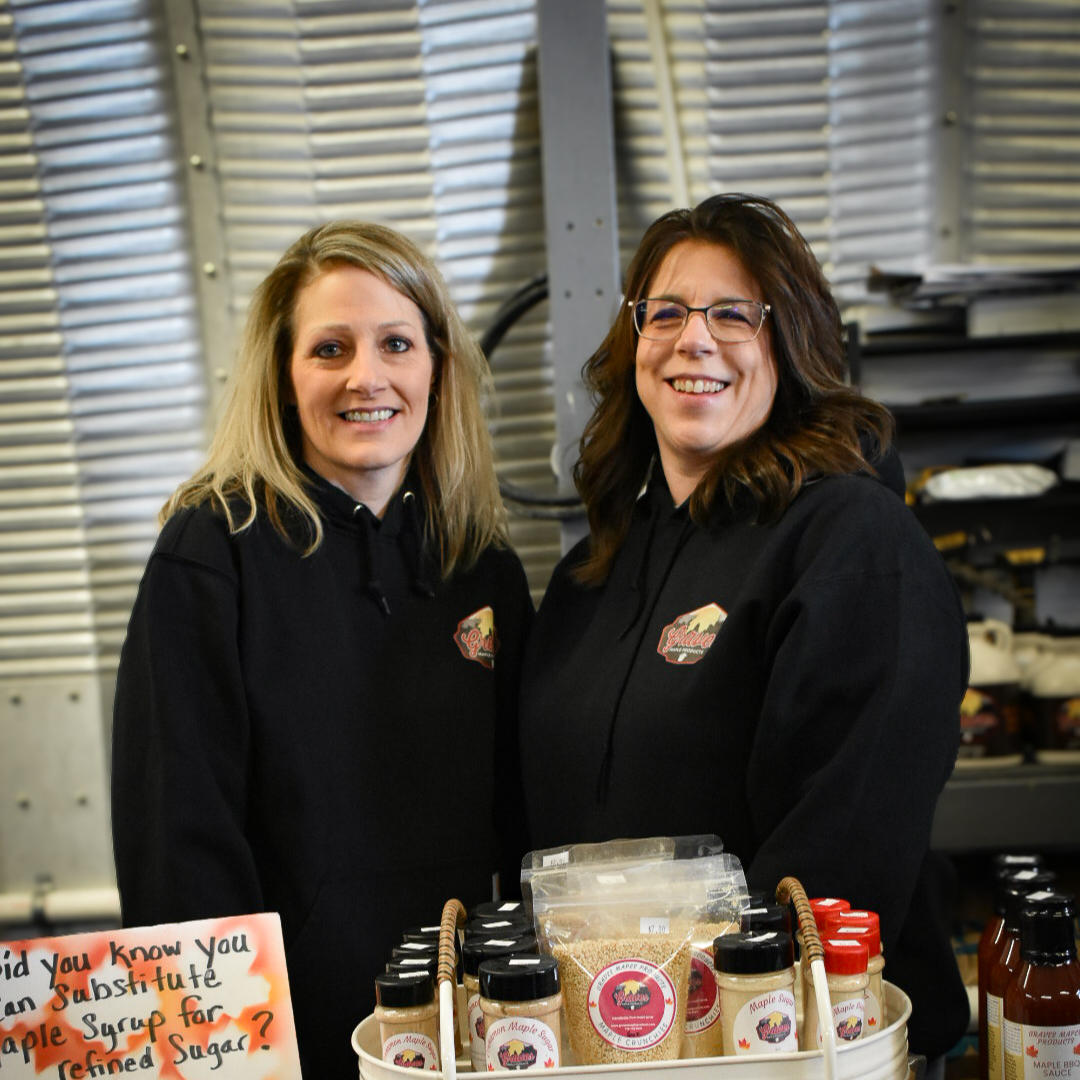 Graves Maple Products team members standing behind a maple gift basket filled with syrup, maple candy, and other handcrafted products, displayed in the sugarhouse.