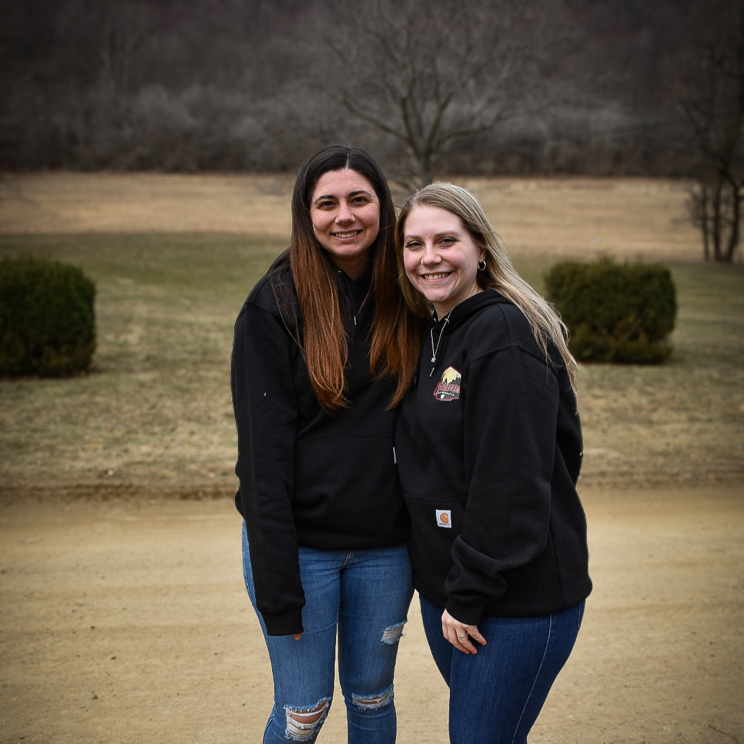 Two Graves Maple Products employees standing outdoors in a grassy field with fall foliage and trees in the background during maple syrup season.
