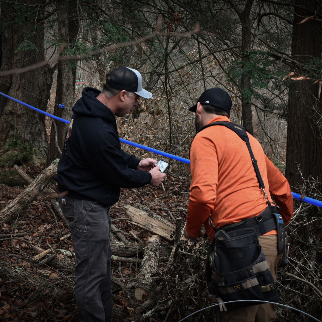 Graves Maple Products Team Reviewing Sap Line Data in the Woods
Two maple syrup workers stand in a forested sugarbush reviewing data on a phone while inspecting a blue maple sap collection line stretched between trees during early-season setup.