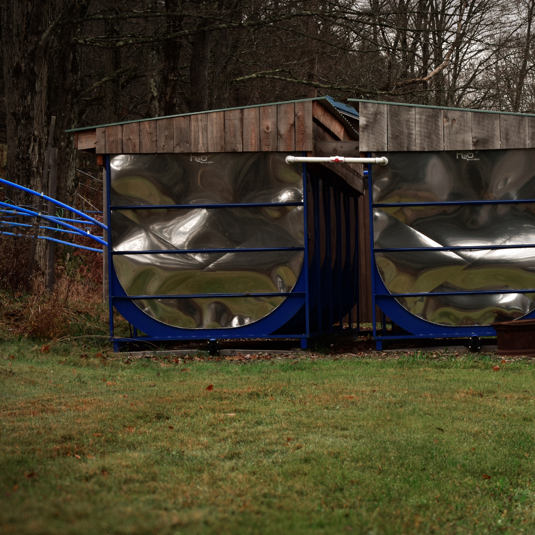 Dual Stainless Steel Sap Tanks for Maple Syrup Production – Graves Maple Products
Two large mirrored sap tanks housed in a wooden shelter collect maple sap through a series of bright blue collection tubes, set against a quiet forest backdrop on the maple farm.